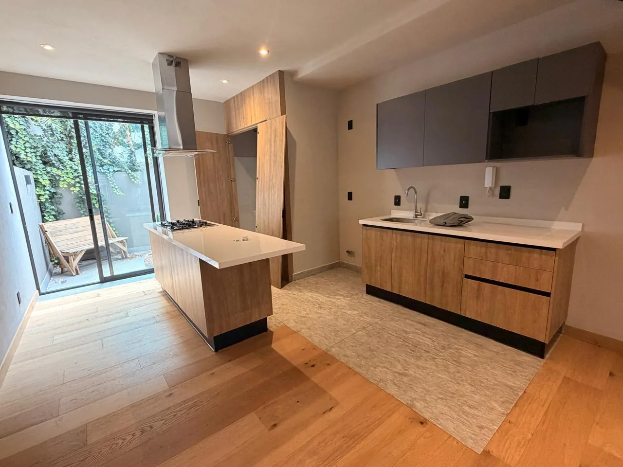 Photo of the real kitchen: an island with a cooktop under a steel hood, a wall counter with a sink, and a glazed door onto a planted terrace.